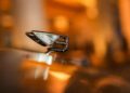 Close-up of a metallic Bentley hood ornament in the shape of a winged "B" on a car, with a blurred warm-toned background—an iconic detail from Bentley Motors' Bespoke Sedans.