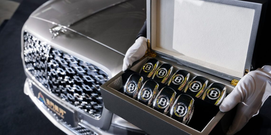 A person wearing white gloves holds an open box containing several Bentley-branded glass tumblers in front of a parked Bentley Motors Bespoke Sedan.