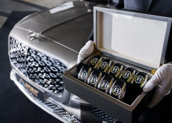 A person wearing white gloves holds an open box containing several Bentley-branded glass tumblers in front of a parked Bentley Motors Bespoke Sedan.