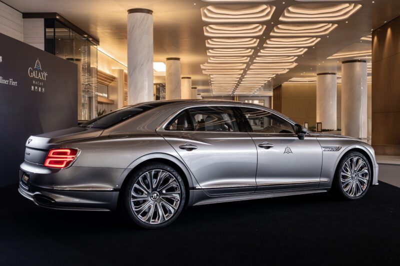 A silver Flying Spur Mulliner luxury sedan from Bentley Motors is displayed indoors on a black platform, featuring distinctive alloy wheels and illuminated ceiling panels above.