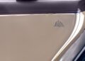 Close-up of a beige car door panel with a black armrest and a small emblem resembling a stylized leaf, reflecting the exquisite craftsmanship found in Bentley Motors’ bespoke sedans like the Flying Spur Mulliner.