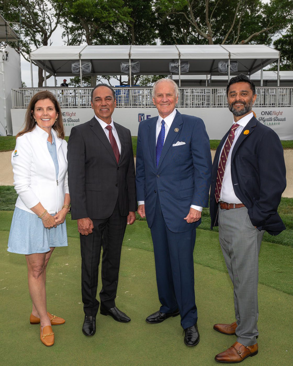 Four people in business attire pose together outdoors on a golf course with event stands and trees in the background.