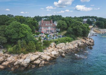 A large house on a rocky shoreline surrounded by trees, with people gathered on the lawn for the Misselwood Concours d’Elegance 15th Anniversary in July and the sea in the foreground under a partly cloudy sky.
