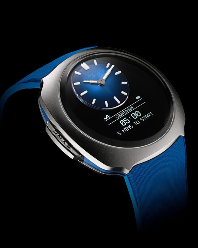 A modern wristwatch with a blue strap, silver bezel, analog clock face, and a digital countdown display reading "05:00 5 MINS TO START," inspired by the precision of Alpine F1 watches.