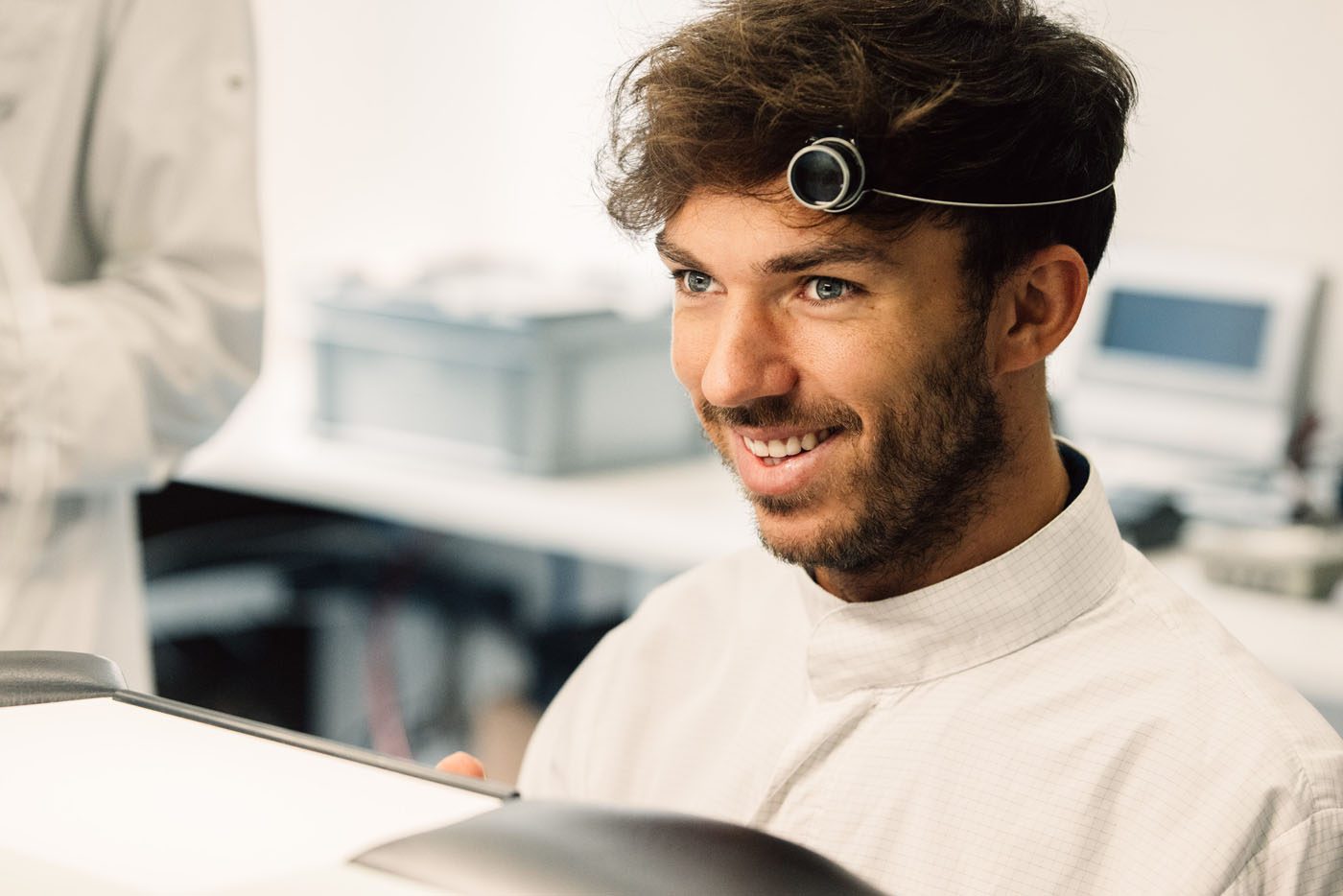 A man wearing a white lab coat and a magnifying loupe on his head smiles while working at a desk in a laboratory, expertly examining an H. Moser & Cie timepiece.
