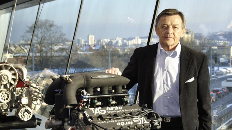 An older man in a suit stands indoors beside a TAG Turbo Porsche engine—the legacy behind the last production Porsche with a Mezger flat-six engine, the GT3 RS 4.0—while city buildings shine through large windows in the background.