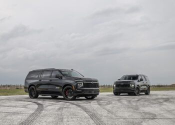 Two black SUVs, including a Hennessey-tuned Chevrolet Tahoe, are parked on an open pavement under a cloudy sky, grassy fields stretching in the background—a true Suburban Upgrade.