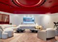 Modern lounge with white and beige seating, a large TV displaying a Ferrari race car, a striped rug, and overhead red ceiling accents. A bar-height table with chairs reflects the sleek Dealer Corporate Identity in the background.