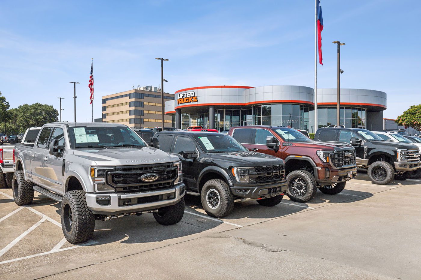 A row of lifted pickup trucks is parked outside America’s Original Lifted Truck Dealer on a clear day.