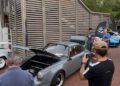 People gather around classic cars on display outdoors at the Celebration Exotic Car Festival; one gray car with its front trunk open is being photographed by a man in the foreground.