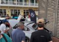 A group of people view classic cars on display outdoors near a yellow building, some hoods open and a black event flag visible at the Celebration Exotic Car Festival.