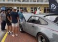 A group of people stands on a sidewalk looking at a gray classic car with its hood open at the Celebration Exotic Car Festival, an outdoor exotic car festival hosted by Classic 9 Motorwerks.