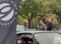 Three men stand next to a car under a Classic 9 Motorwerks banner, admiring the vehicle’s interior at the Celebration Exotic Car Festival, with trees in the background at this outdoor event.