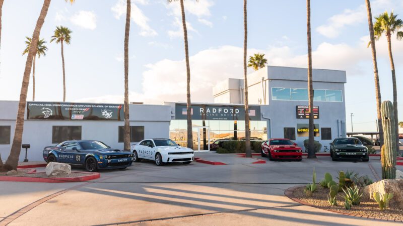 Four cars are parked in front of the Radford Racing School building under a partly cloudy sky, with palm trees and desert plants visible around the entrance.