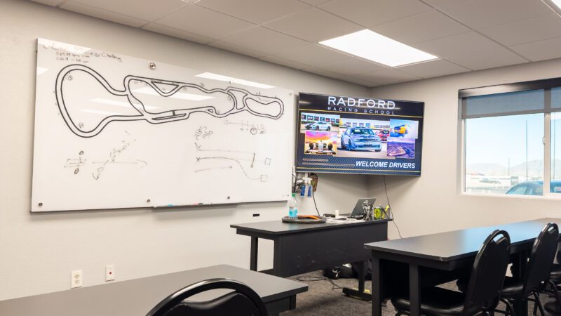 Classroom with a whiteboard showing a racetrack diagram, a desk, and a screen displaying "Radford Racing School" and "Welcome Drivers.