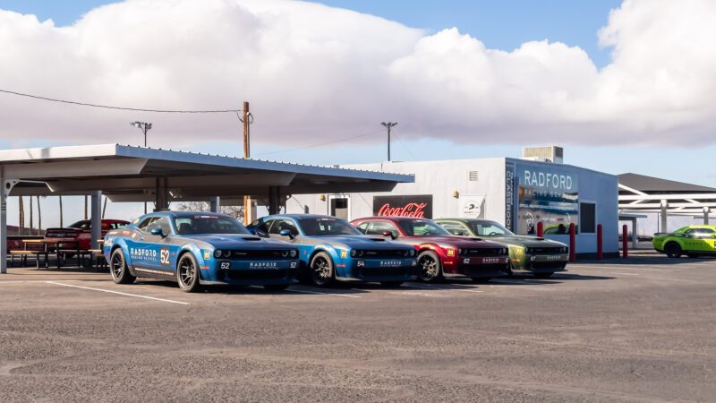 Several Dodge sports cars are parked in front of a Radford Driving School building on a sunny day.