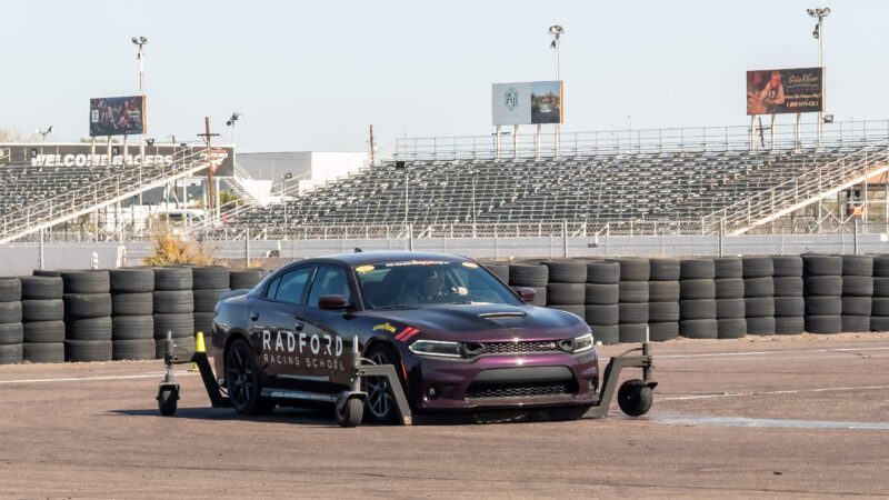 A maroon car labeled "Radford Racing School" is equipped with training outriggers on a racetrack, with empty grandstands and billboards in the background.