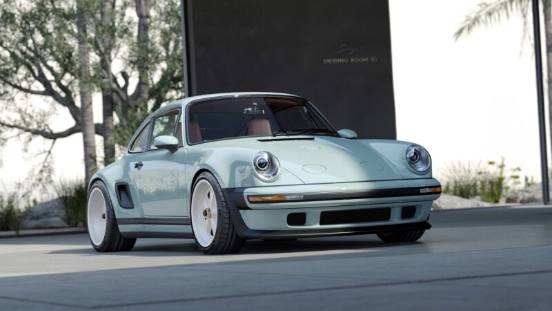 A light blue, limited edition vintage-style Porsche 911 by Singer with round headlights and wide tires is parked outdoors near a building with trees in the background.