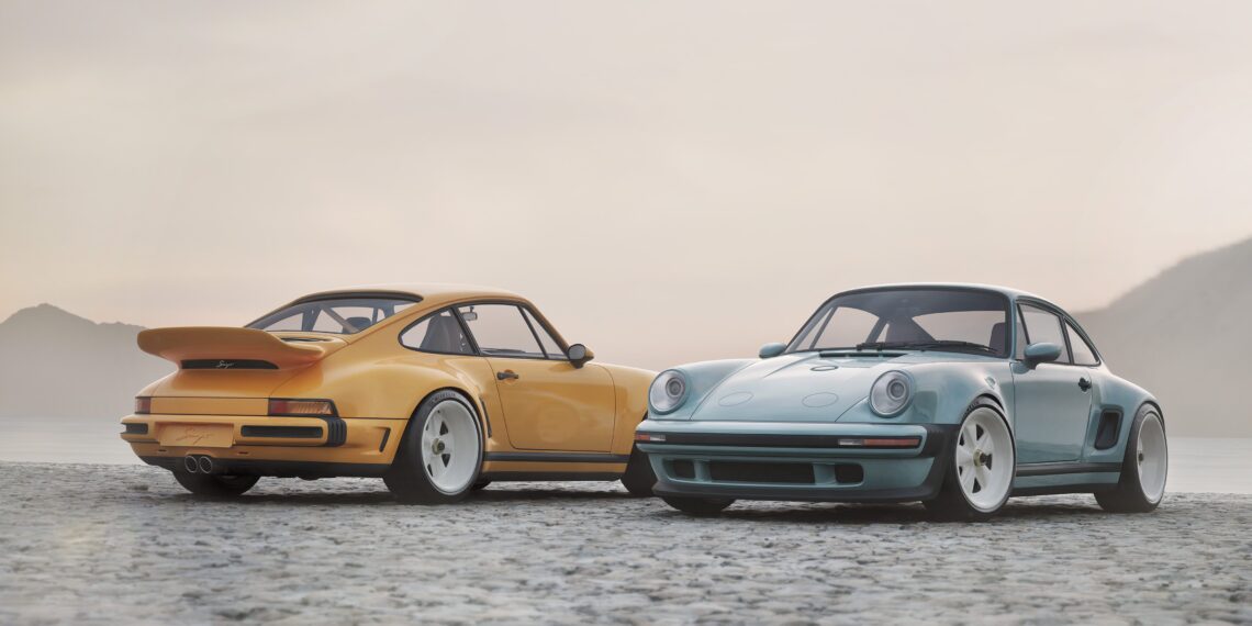 Two vintage sports cars, including a limited edition Porsche 911 by Singer in yellow and a classic blue model, are parked side by side on a cobblestone surface near a misty lake with mountains in the background.