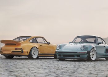 Two vintage sports cars, including a limited edition Porsche 911 by Singer in yellow and a classic blue model, are parked side by side on a cobblestone surface near a misty lake with mountains in the background.