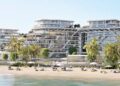 Modern beachfront resort with multiple glass-fronted buildings, palm trees, lounge chairs, and umbrellas along a sandy shore—part of the stunning $1.3B Jacob & Co. beachfront development announced south of Dubai. The sky is clear and the water is calm.