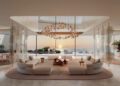 A modern, spacious living room with large windows overlooking the ocean at sunset, furnished with white sofas, a chandelier, and indoor plants—evoking the luxury of Jacob & Co.’s $1.3B beachfront development south of Dubai.