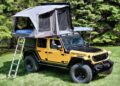 A yellow Jeep with large tires is parked on grass and equipped with a carbon-fiber rooftop tent—a game changer for the Jeep Wrangler—accessible by a metal ladder and surrounded by green trees.
