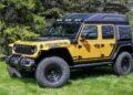 A yellow and black Jeep Wrangler with large off-road tires and a raised camper-style carbon-fiber tent—one thing that makes this a game changer for the Jeep Wrangler—is parked on grass near trees.