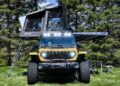 A yellow off-road vehicle with a roof-top tent—a true game changer for the Jeep Wrangler thanks to its carbon-fiber build—is parked on grass near folding chairs and a table, surrounded by pine trees.