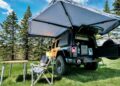A yellow off-road vehicle with an open rooftop tent—one thing that makes this carbon-fiber tent a game changer for the Jeep Wrangler—is parked on grass beside folding chairs and a table, with pine trees behind under a blue sky.