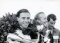 Three men, two wearing laurel wreaths and one in a racing suit, smile and stand together after a motor racing event featuring the exclusive 1-of-60 Emira Jim Clark Edition Lotus.