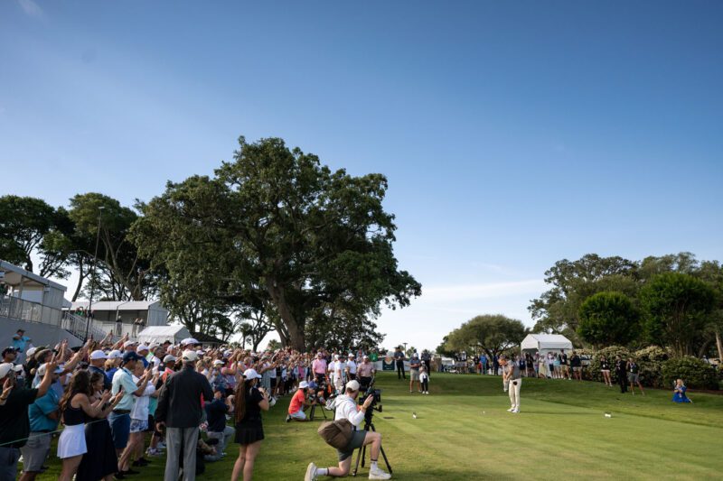 A crowd watches a golfer preparing to take a shot on a green course under a clear blue sky, with trees and tents in the background.