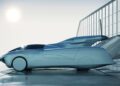 A sleek, futuristic car—resembling the production prototype of Klein Vision AirCar—rests by a metal fence on a reflective surface, its large rear wing gleaming as the sun shines in the background.