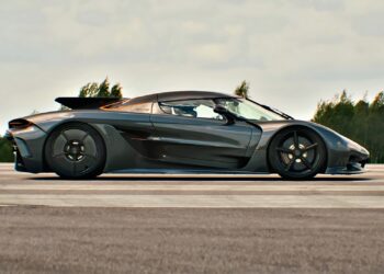 A sleek black sports car, reminiscent of the Koenigsegg Jesko Absolut, is parked on a road lined with trees under a cloudy sky, exuding power worthy of the 1/2 Mile Top Speed Record.