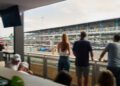 Spectators watch a blue Formula 1 car race past from a covered viewing area overlooking the track and grandstands.