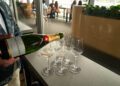 A person pours Moët & Chandon champagne into a glass next to three empty wine glasses on a table by a window; people sit and talk in the background.