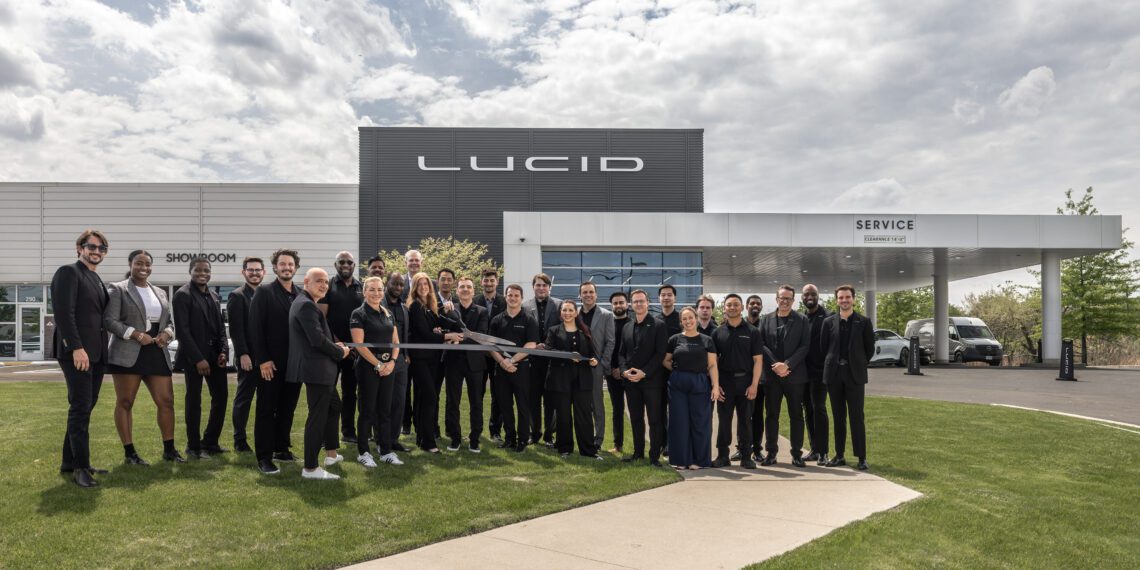 A group of people poses in front of a Lucid building and service center, with several individuals holding a large ceremonial ribbon-cutting scissors.