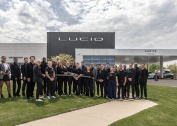 A group of people poses in front of a Lucid building and service center, with several individuals holding a large ceremonial ribbon-cutting scissors.