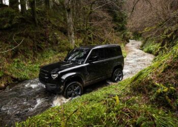 A black SUV drives through a shallow stream surrounded by green grass and trees in a forested area during the Destination Defender Festival in New York’s scenic Hudson Valley.