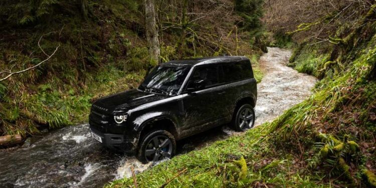 A black SUV drives through a shallow stream surrounded by green grass and trees in a forested area during the Destination Defender Festival in New York’s scenic Hudson Valley.