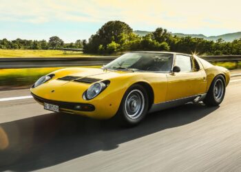 A yellow Lamborghini, celebrated in automotive history and restored by Polo Storico, drives on a highway with trees and mountains in the background on a sunny day.