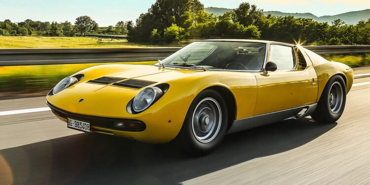 A yellow Lamborghini, celebrated in automotive history and restored by Polo Storico, drives on a highway with trees and mountains in the background on a sunny day.