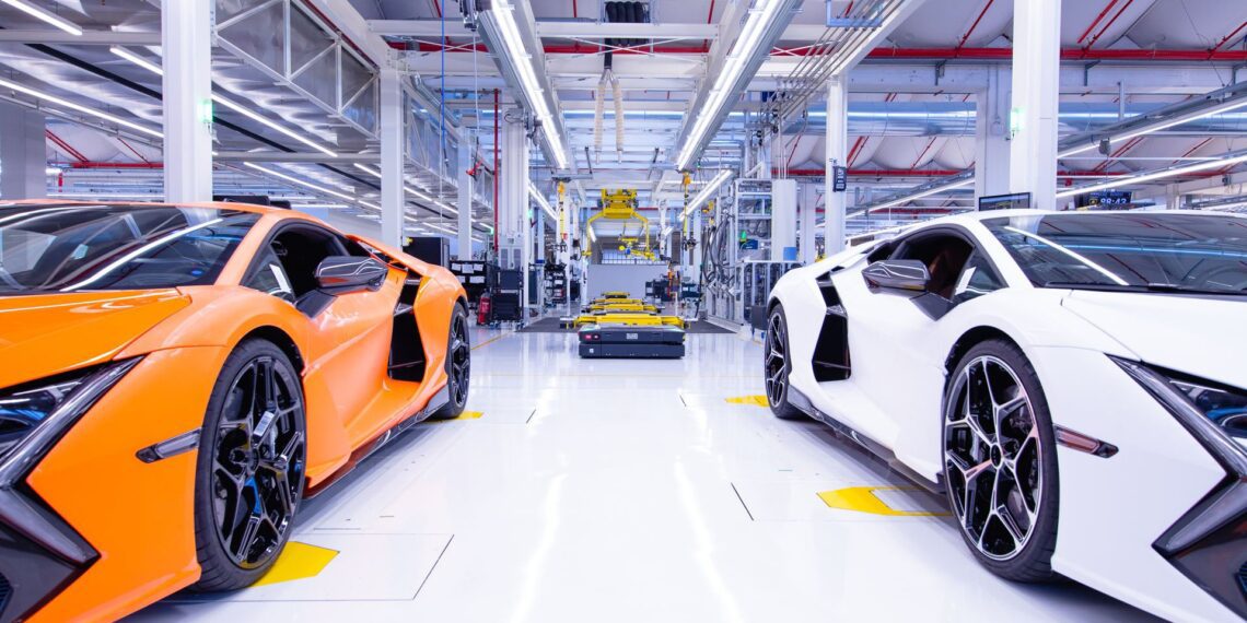 Orange and white sports cars are parked inside a bright, modern automotive manufacturing facility, reflecting the innovation behind Automobili Lamborghini Continues To See Growth In Q1 2025. Machinery and equipment fill the background.