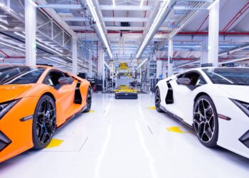 Orange and white sports cars are parked inside a bright, modern automotive manufacturing facility, reflecting the innovation behind Automobili Lamborghini Continues To See Growth In Q1 2025. Machinery and equipment fill the background.