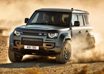 A dark gray Land Rover Defender, a symbol of Here's What's Driving JLR's Best Annual Profits In Nearly A Decade, speeds along a dusty off-road trail, kicking up dirt clouds with its driver clearly visible inside.