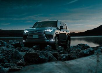 A Lexus SUV, embodying Expedition Elegance, sits with headlights aglow on rocky terrain by a lake at dusk, framed by hills and a darkening sky.