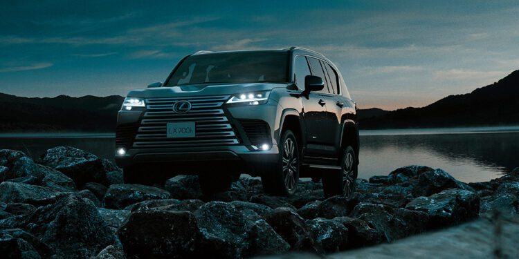 A Lexus SUV, embodying Expedition Elegance, sits with headlights aglow on rocky terrain by a lake at dusk, framed by hills and a darkening sky.