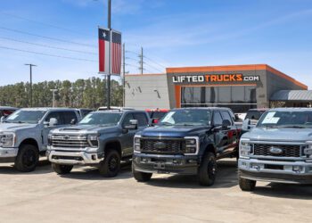 Four large pickup trucks are parked in front of a dealership with a "LIFTEDTRUCKS.com" sign, proudly showcasing America’s Original Lifted Truck Dealer, with a Texas flag visible in the background.