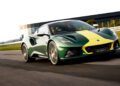 A green Lotus Emira Jim Clark Edition, 1-of-60, with a yellow racing stripe drives on a racetrack, with a modern building and grass in the background.