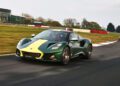 A green and yellow Lotus Emira Jim Clark Edition, 1-of-60, speeds along a racetrack with a building and trees in the background.